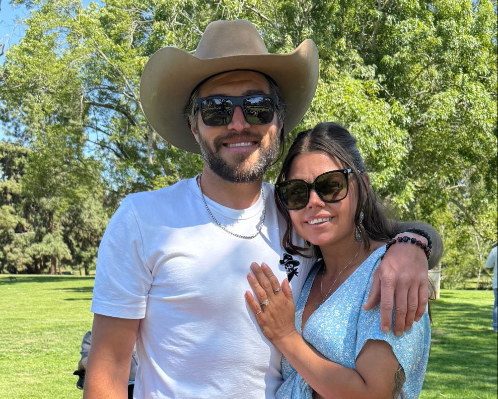 Man and woman posing on a red blanket in a park with trees and grass in the background.